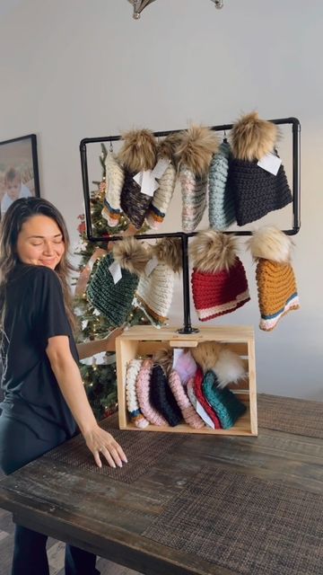 a woman standing next to a wooden table in front of a display of hats and mittens