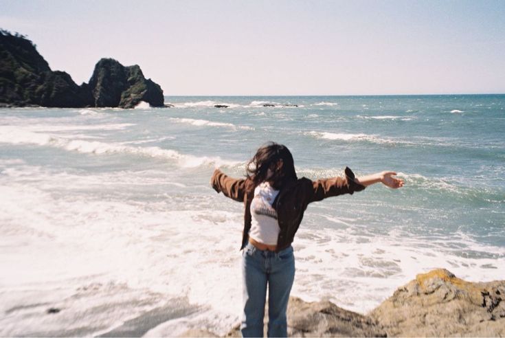 a woman standing on top of a rocky beach next to the ocean with her arms outstretched