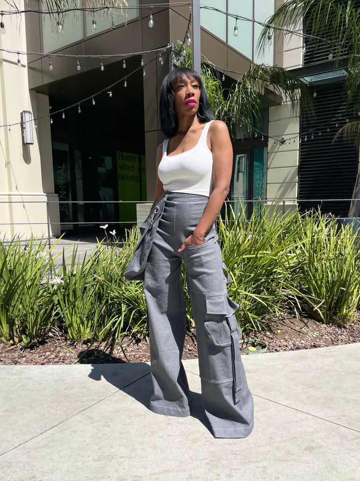 a woman standing on the sidewalk in front of a building wearing grey pants and a white tank top