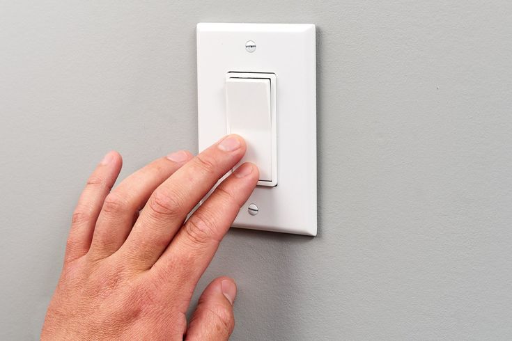 a hand is pressing the light switch button on a white wall mounted electrical outlet with grey walls