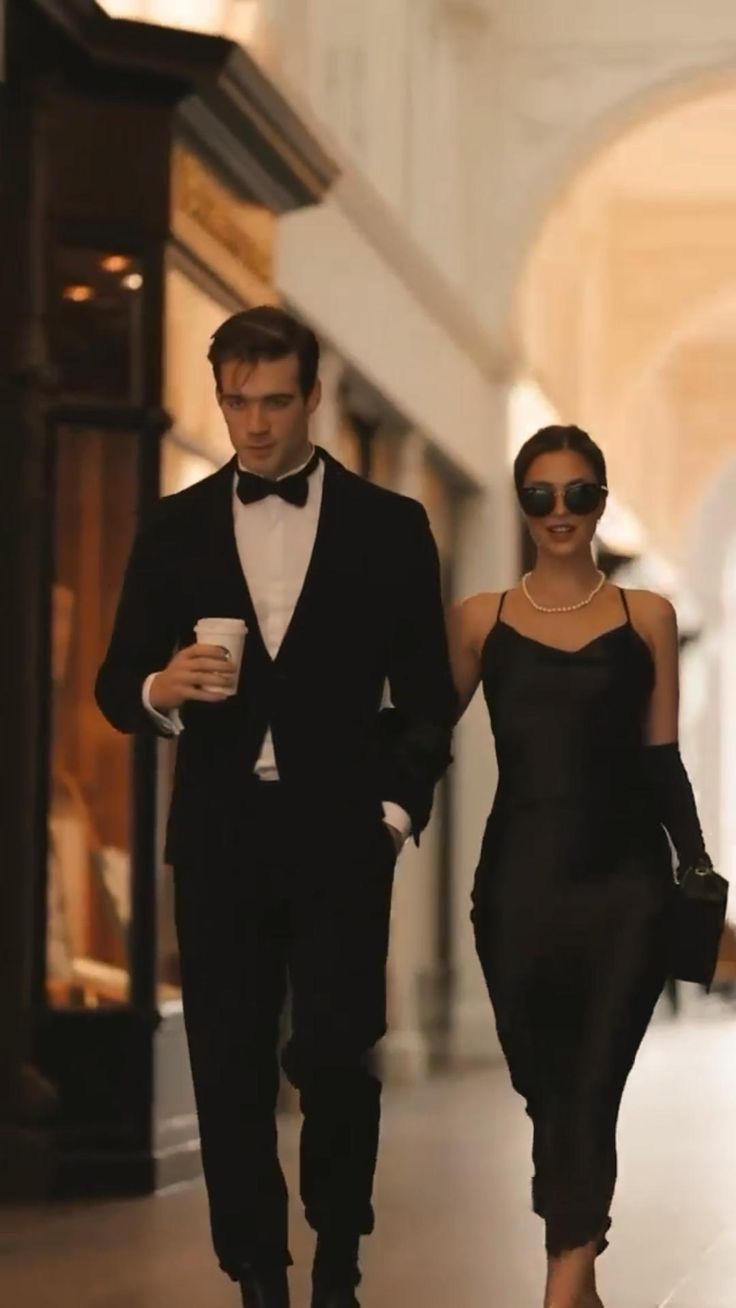 a man and woman dressed in black walking down the street with one holding a coffee cup