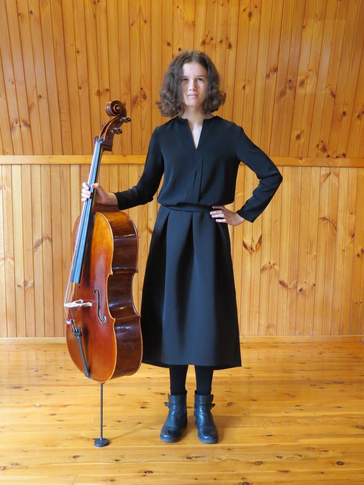 a woman standing next to a cello on top of a hard wood floor in front of a wooden wall