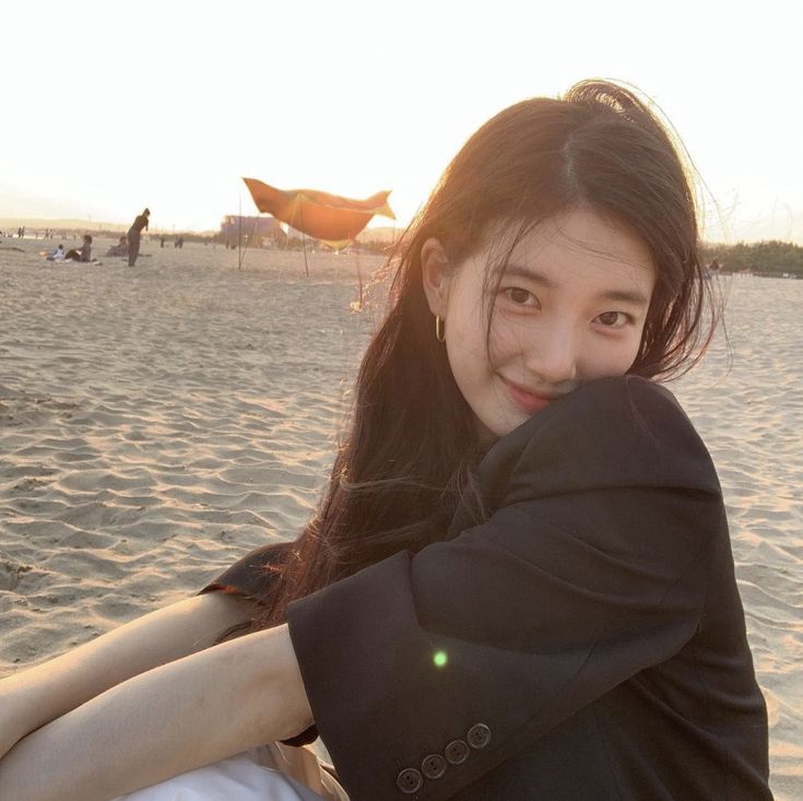 a woman sitting on top of a sandy beach next to the ocean with her arms around her neck