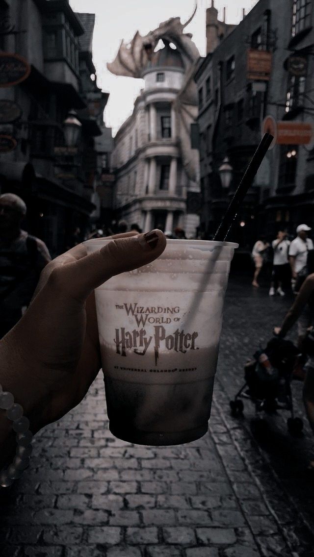 a hand holding up a drink in the middle of a brick street with people walking around