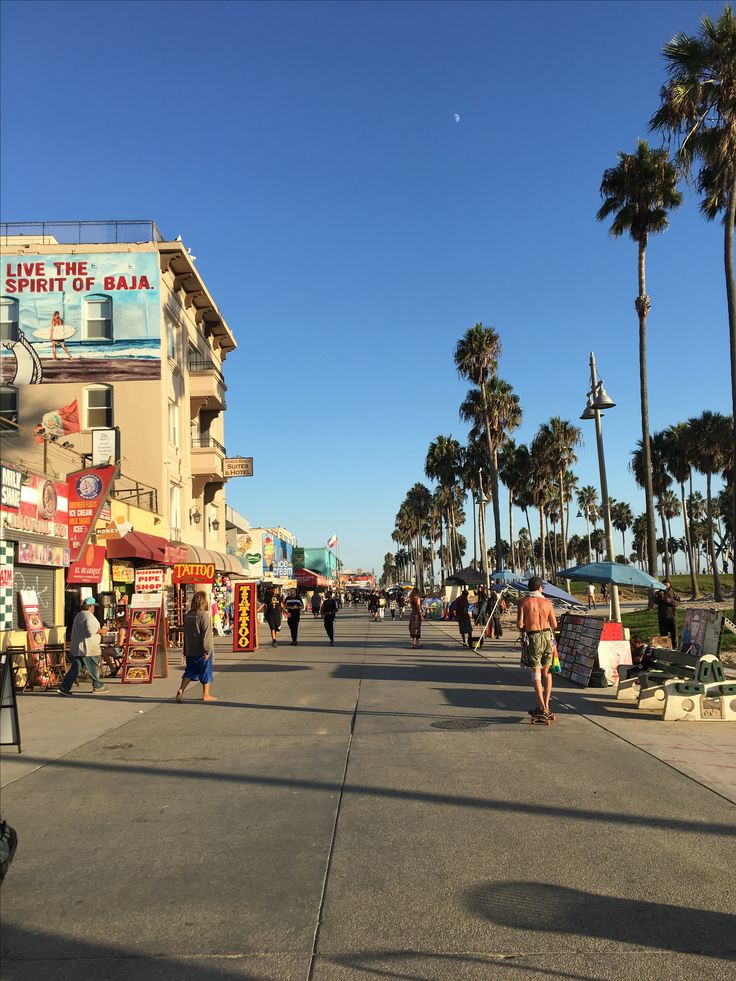 people are walking down the street in front of shops and palm trees on a sunny day