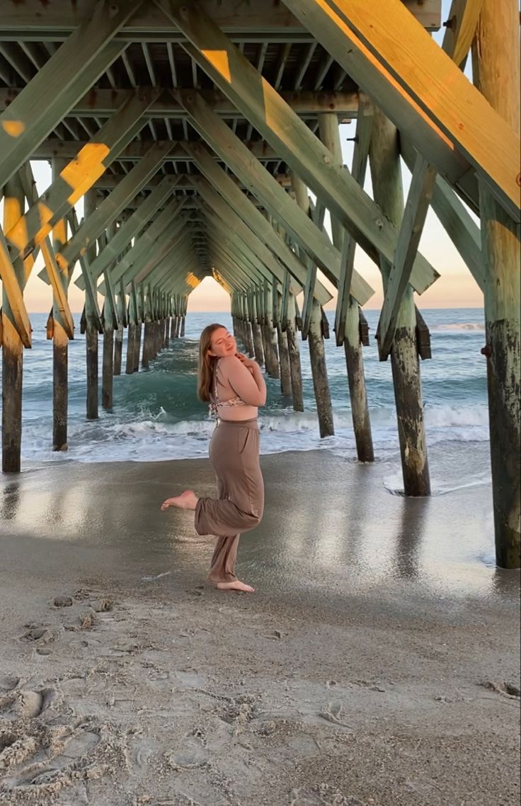 a woman standing under a wooden pier next to the ocean with her legs in the air