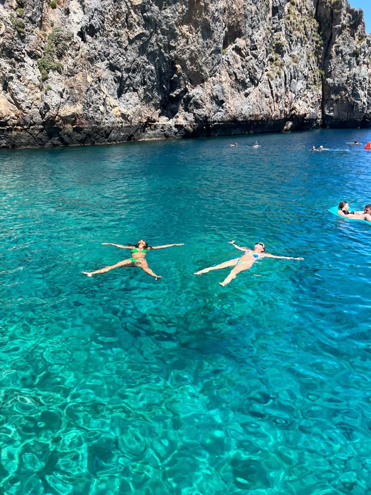 three people swimming in clear blue water next to cliffs