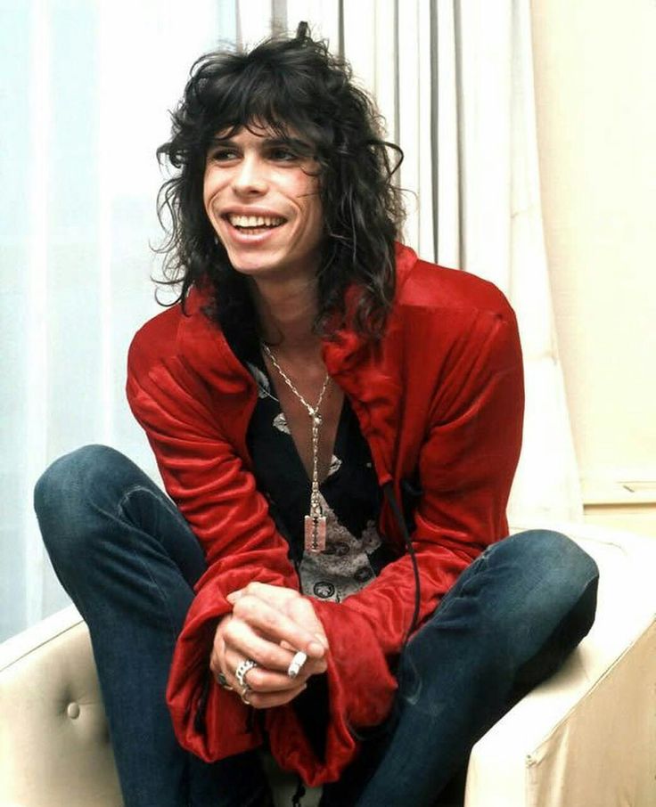 a young man with long hair sitting on the floor smiling at the camera while wearing blue jeans and a red shirt