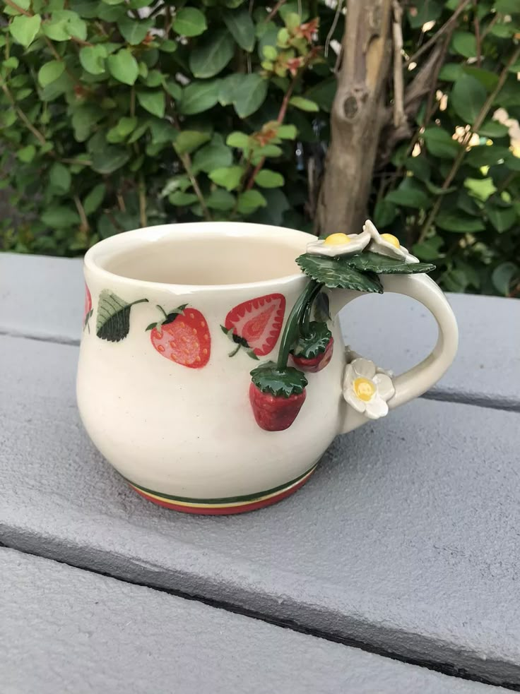 a cup with strawberries painted on it sitting on a table next to some bushes