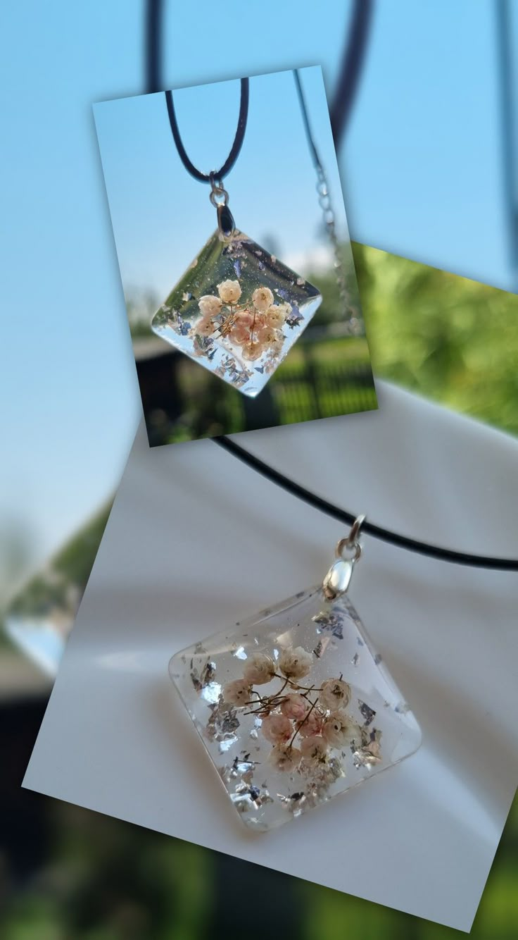 two square pendants with flowers on them hanging from a black leather cord and an open window in the background