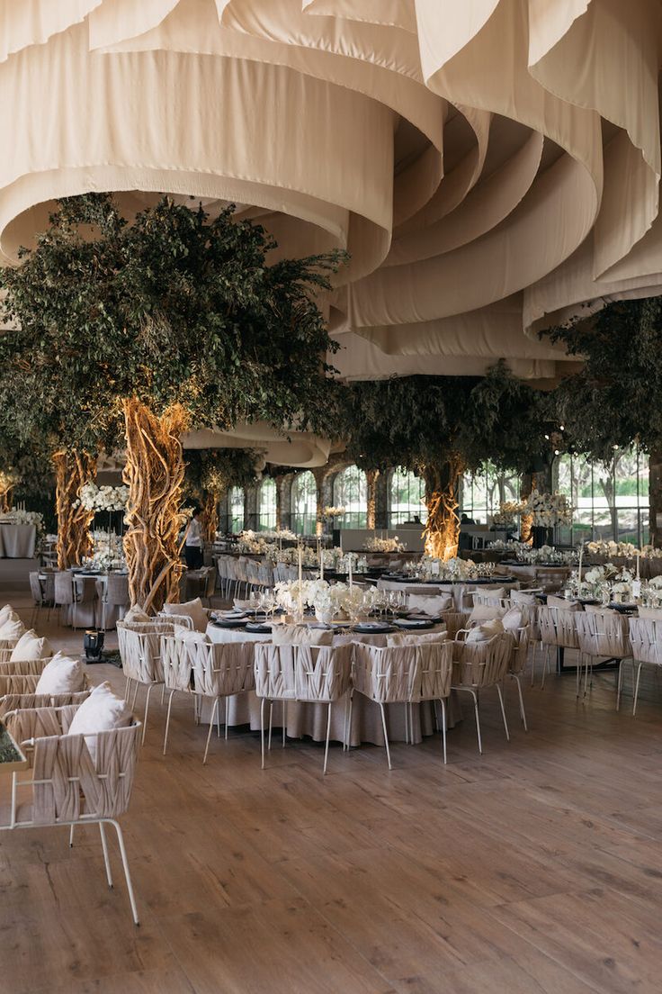 a large room with tables and chairs covered in white linens, trees and hanging lights