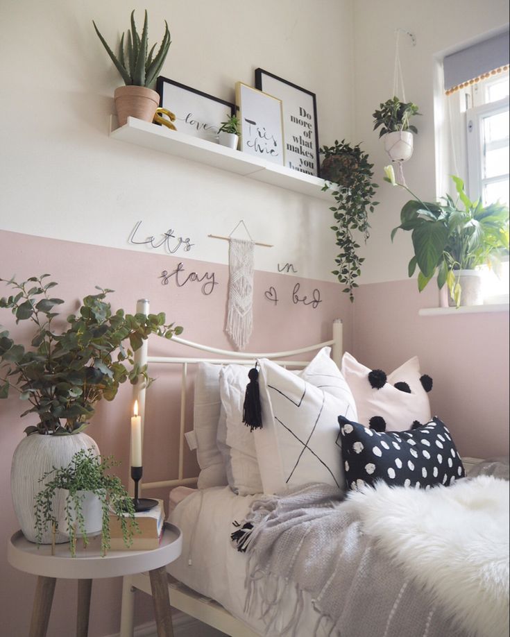 a white bed topped with lots of pillows next to a window filled with potted plants