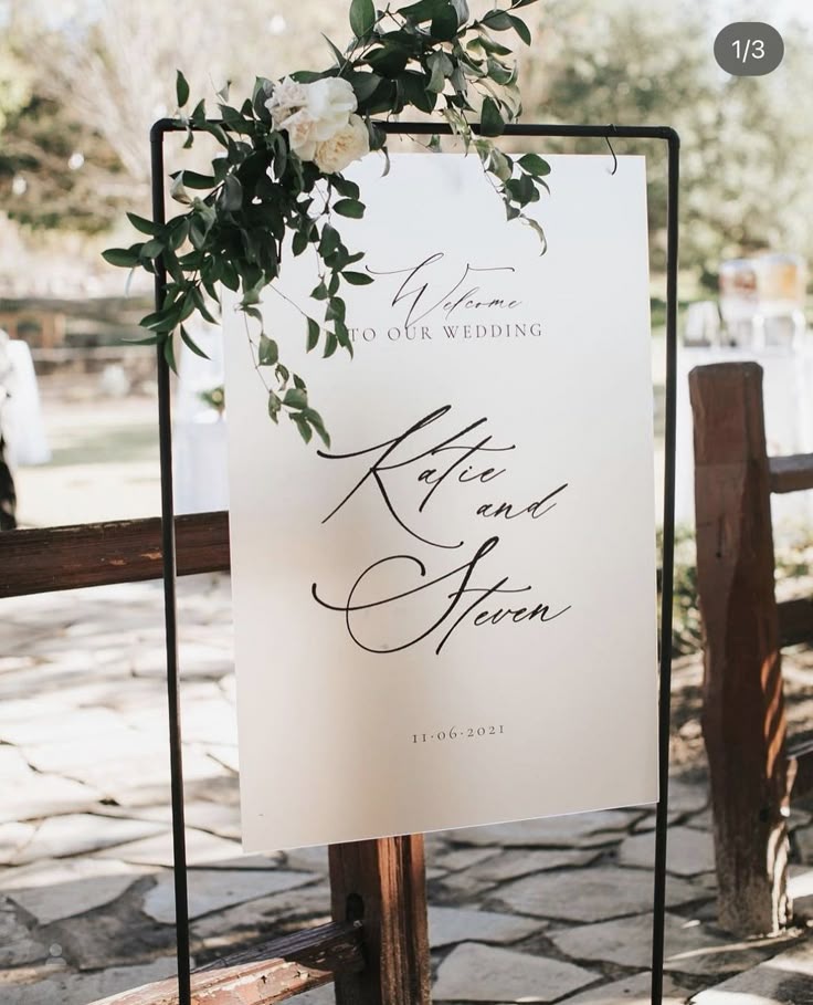 a wedding sign with flowers on it sitting next to a wooden fence in front of a stone walkway