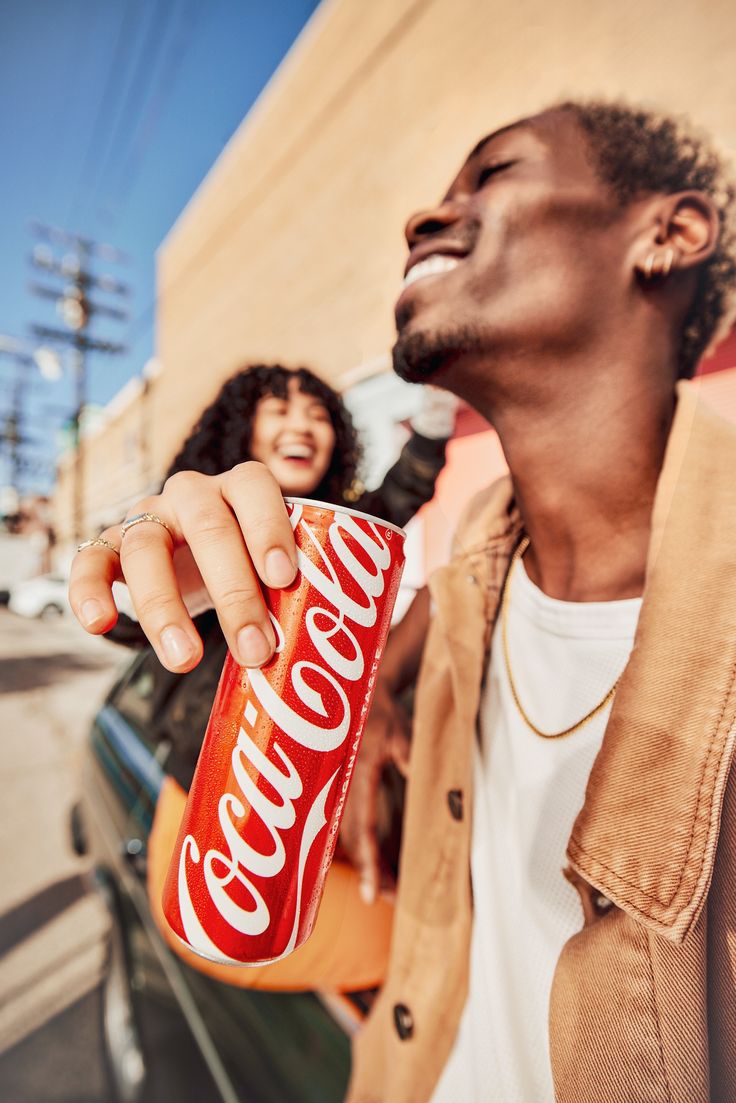 a man holding a can of coca - cola in his right hand while standing next to a woman