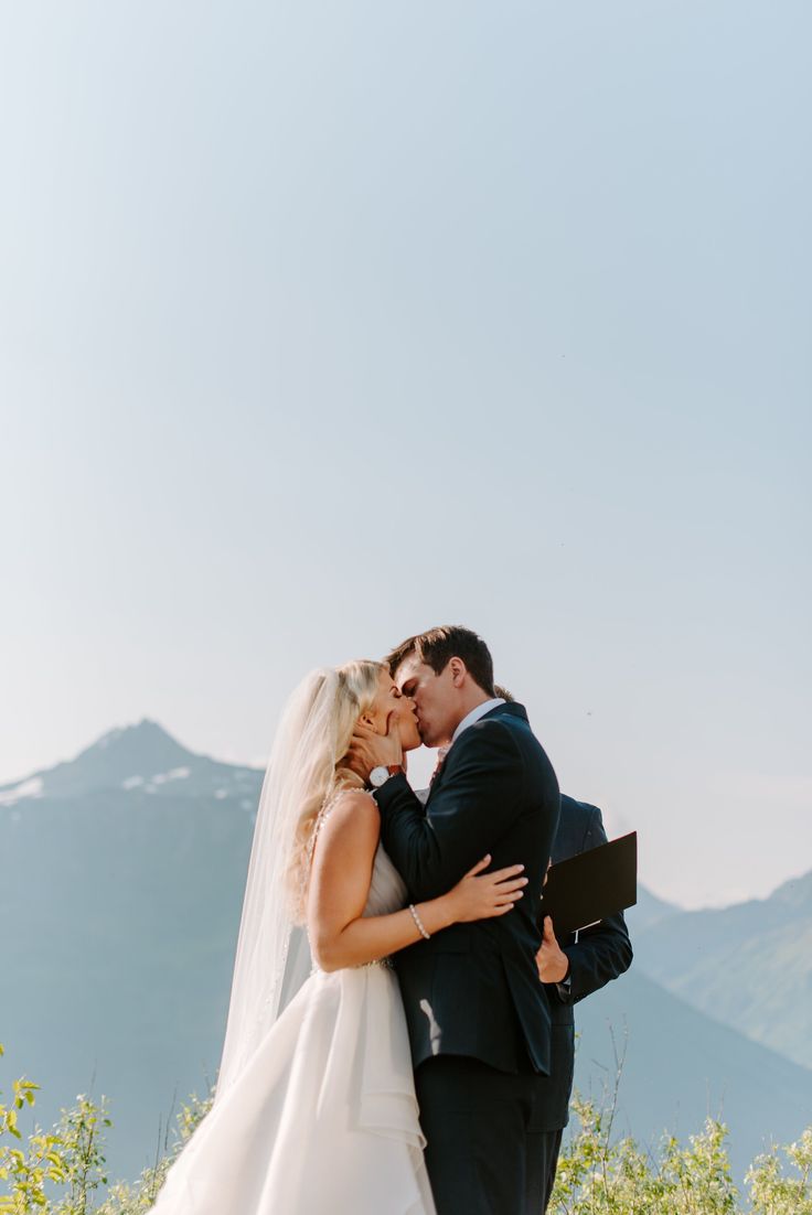 a bride and groom kissing on top of a hill with mountains in the back ground