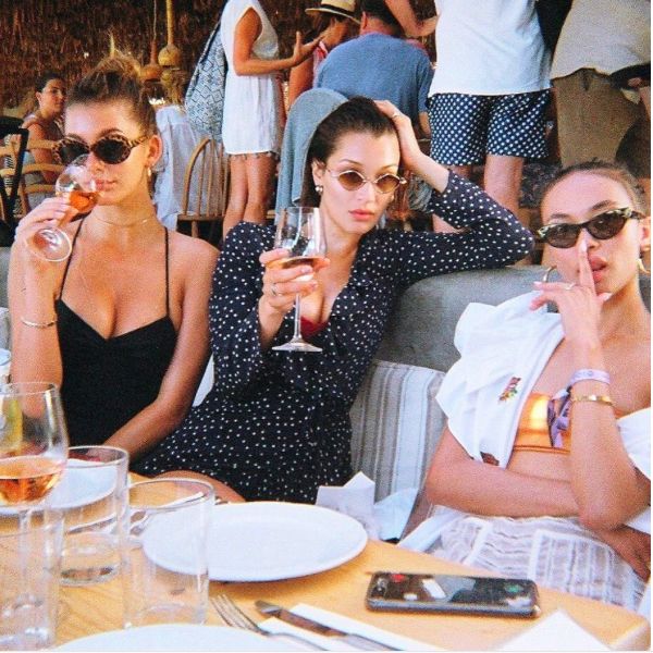 three women sitting at a table with wine glasses in their hands and people standing around them