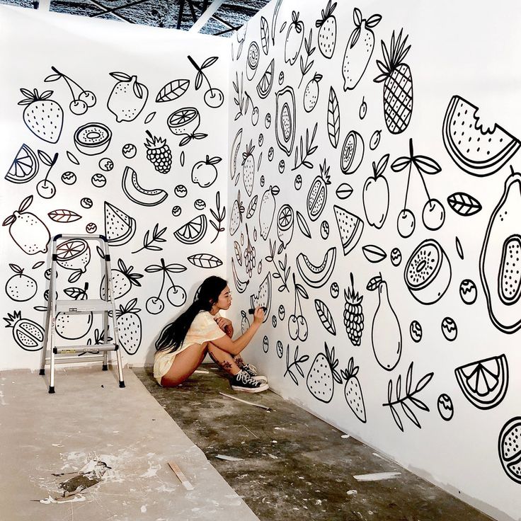 a woman sitting on the ground in front of a wall with fruit drawings painted on it