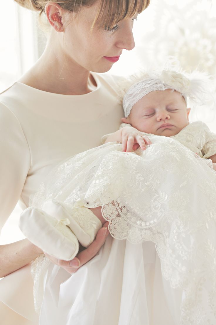 a woman holding a baby wearing a white dress and headband with her hands on her chest