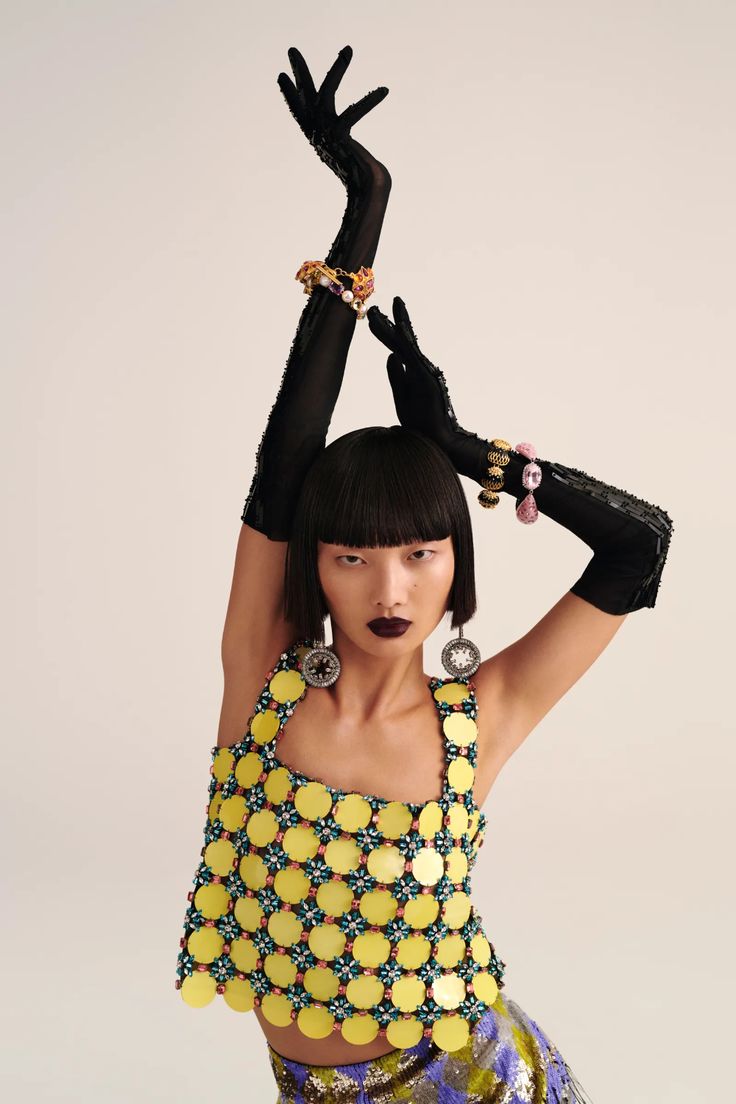 a woman with her hands on her head wearing black gloves and yellow dress, holding an object in the air