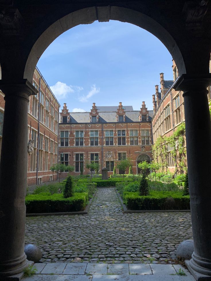 an archway leading to a building with many windows and plants in the courtyard, surrounded by stone walkways