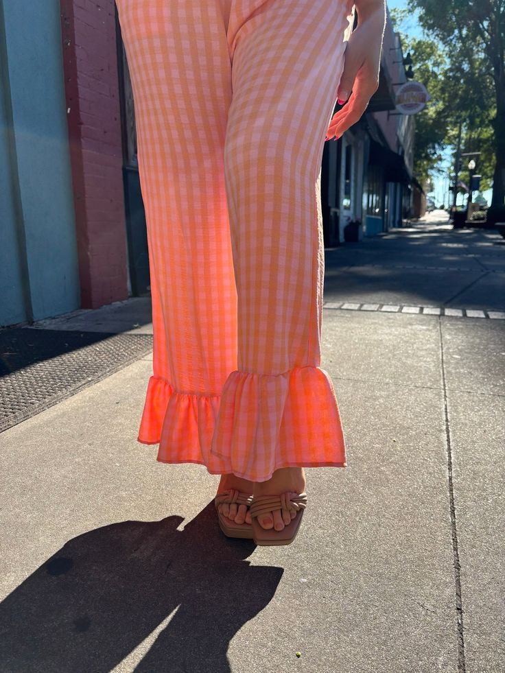 These fun and quirky pants will add some excitement to your wardrobe. The wide leg, elastic waistband, and relaxed fit offer comfort and versatility, while the pink and orange gingham and ruffled hemline add a playful touch. Perfect for dressing up or down, these pants are a dream come true! Chic Summer Pants With Flared Hem, Casual Flare Bottoms With Ruffles, Casual Cotton Bottoms With Flared Hem, Spring Peach Bottoms For Loungewear, Casual Wide-leg Pants With Ruffles, Casual Ruffled Flare Bottoms, Trendy Flared Hem Summer Pants, Casual Spring Wide Leg Pants With Flared Hem, Summer Flare Bottoms With Ruffles