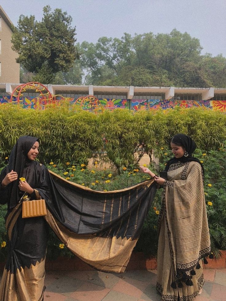 two women standing next to each other in front of some bushes and flowers, one holding a large black tarp