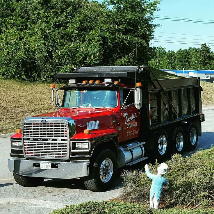 a red dump truck driving down a road next to a lush green field and forest