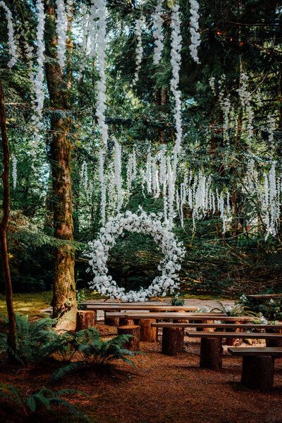 an outdoor wedding ceremony in the woods with white flowers hanging from the trees and wooden benches