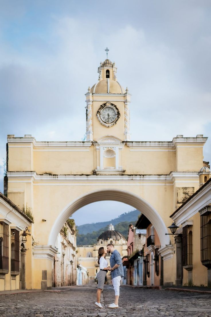 two people standing in front of an archway with a clock on the top of it