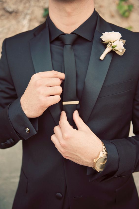 a man in a black suit adjusting his tie with a white flower on the lapel