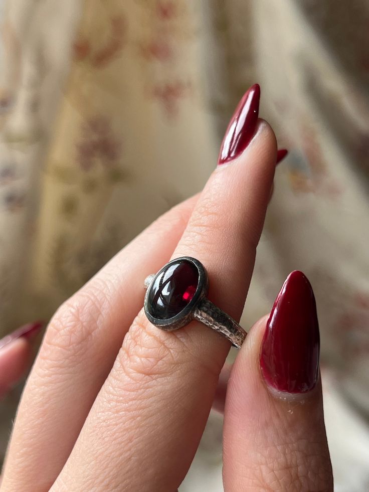 A gorgeous and vampy adornment that is a custom Curio original! These hand-made pieces are a true treasure, for they are one of a kind and crafted with intention to marry the aesthetics of the past, with the modern stylings of the present. This hammered silver garnet ring features a stunning blood-red garnet cabochon set in striking sterling silver. The band is simple and understated, with fine scrolling foliate detail that bring a gentle glamour to the silhouette. There is versatility in this design as well – pair it with a casual or formal look, or even go another step ahead and loop it through a delicate chain as a charm. Either way, this Curio Original is a magnificent addition for any jewelry lover’s collection! Materials and Features: Sterling Silver, Garnet Cabochon Age/Origin: Curi Heirloom Garnet Jewelry For Gifts, Unique Ruby Gemstone Jewelry, Hand Forged Rings As Gift, Handmade Gothic Open Ring, Unique Hallmarked Ruby Ring Gift, Handmade Elegant Ruby Ring, Unique Hallmarked Ruby Ring As A Gift, Elegant Handmade Ruby Ring, Hand Forged Oval Ring Fine Jewelry