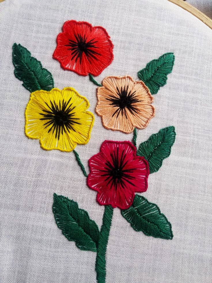 three colorful flowers on a white cloth with green and red leaves are in the center of this embroidery work