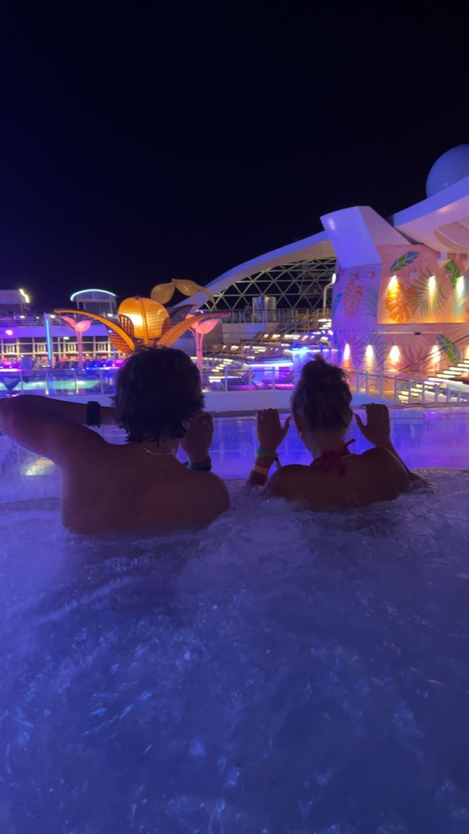 two people sitting in a hot tub at night with lights on the building behind them