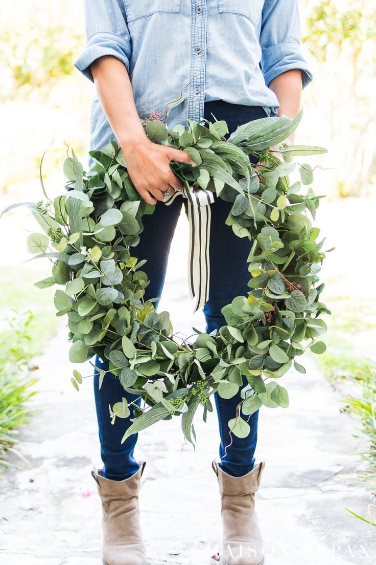 a person holding a wreath with greenery on the front and side, while wearing boots