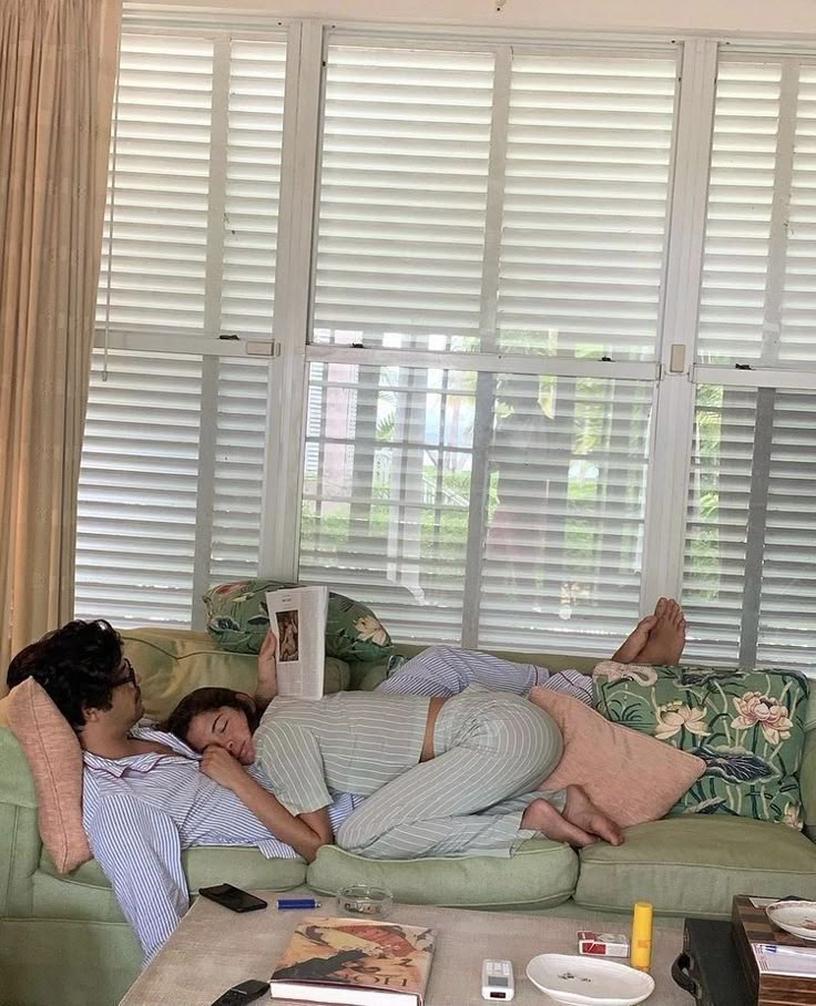 two people laying on couches in front of a window with shutters and blinds