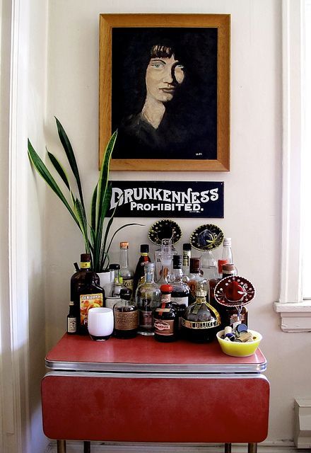 a red table topped with lots of bottles and glasses next to a painting on the wall