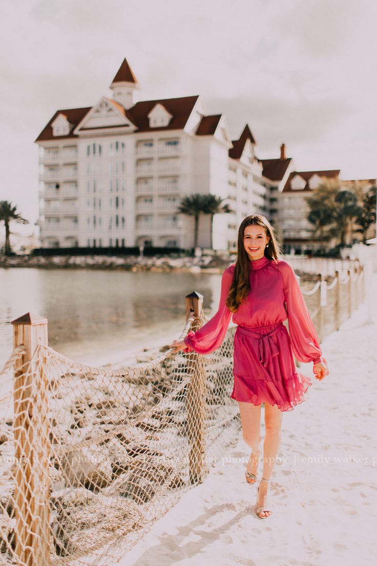 a woman in a pink dress is walking on the beach next to a netted fence