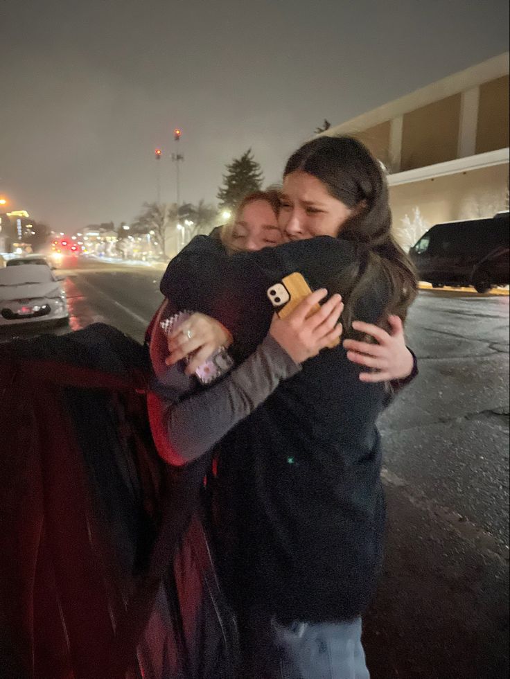 two women hugging each other on the side of the road in front of some cars