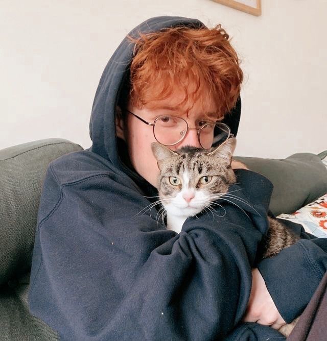 a woman with glasses holding a cat on her lap while wearing a hoodie over her face