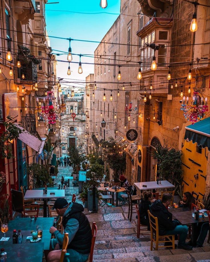 people sitting at tables in an alleyway with lights strung over the buildings on either side