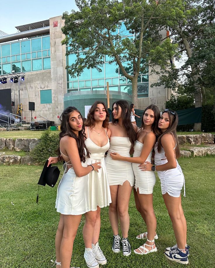 four women in white dresses posing for the camera on grass with trees and building behind them