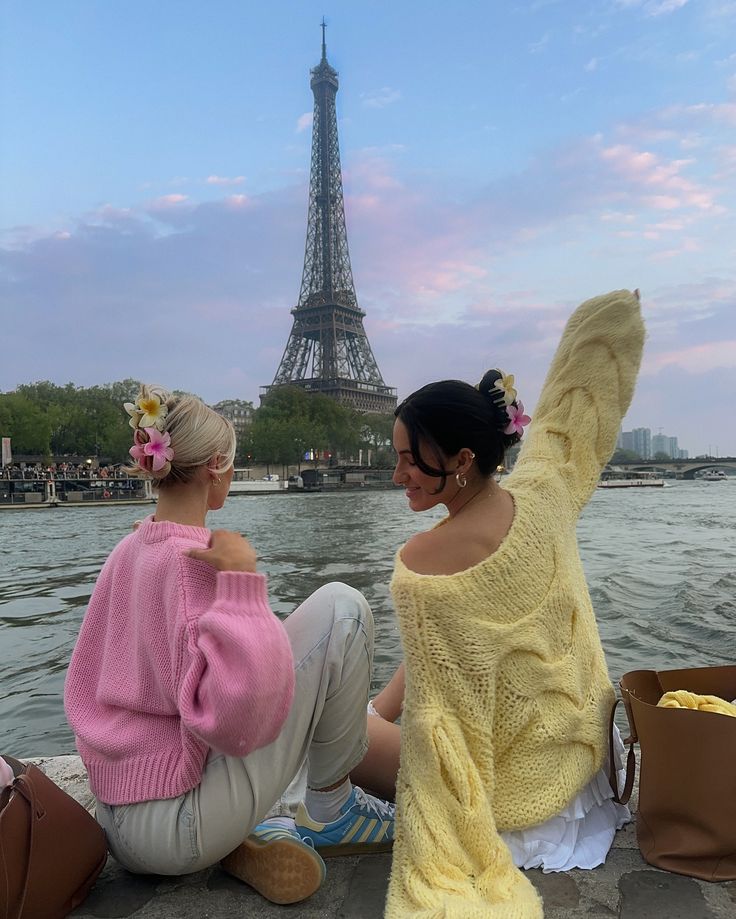 two women sitting next to each other near the water in front of the eiffel tower