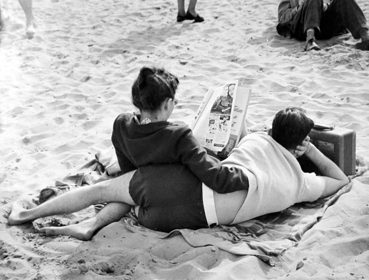 two people laying on the sand reading a book