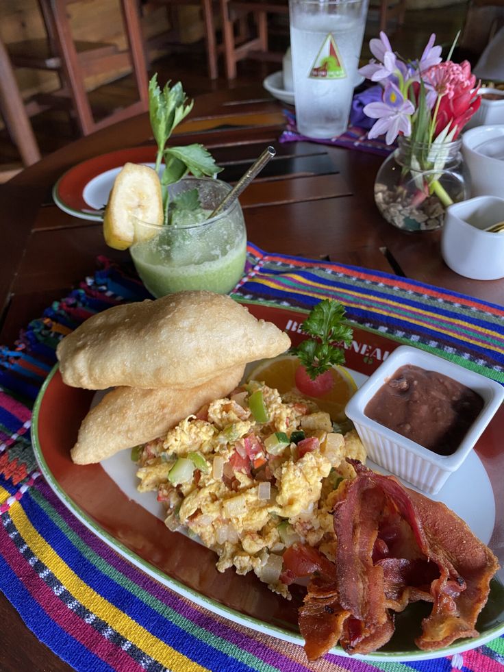 a breakfast plate with eggs, bacon, toast and fruit on it sitting on a colorful table cloth