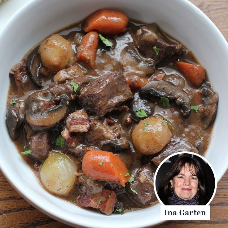 a white bowl filled with stew and carrots on top of a wooden table next to a woman's face