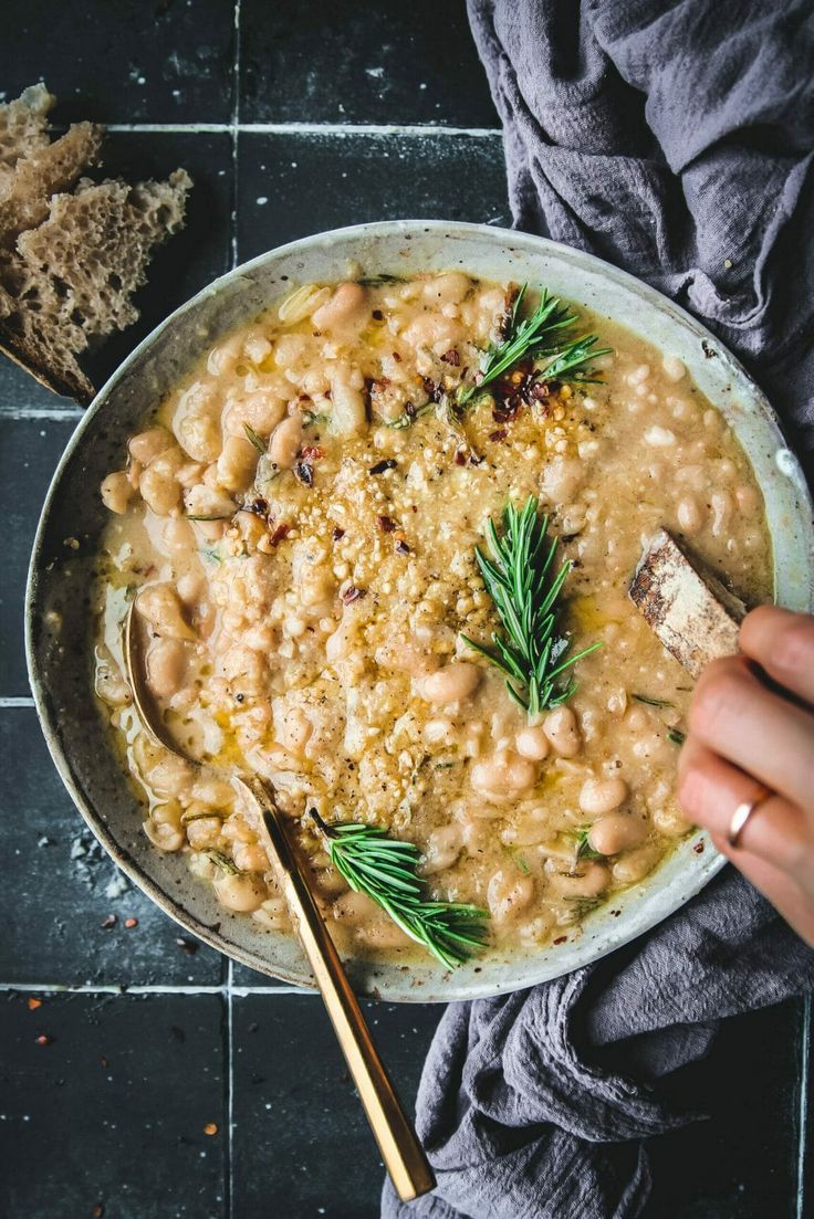 a person is holding a spoon in a bowl of beans and herbs with bread on the side