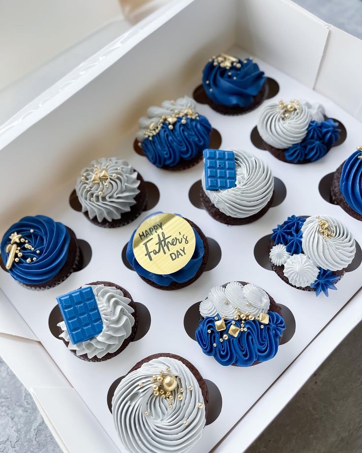 twelve decorated cupcakes in a white box with blue frosting and gold decorations