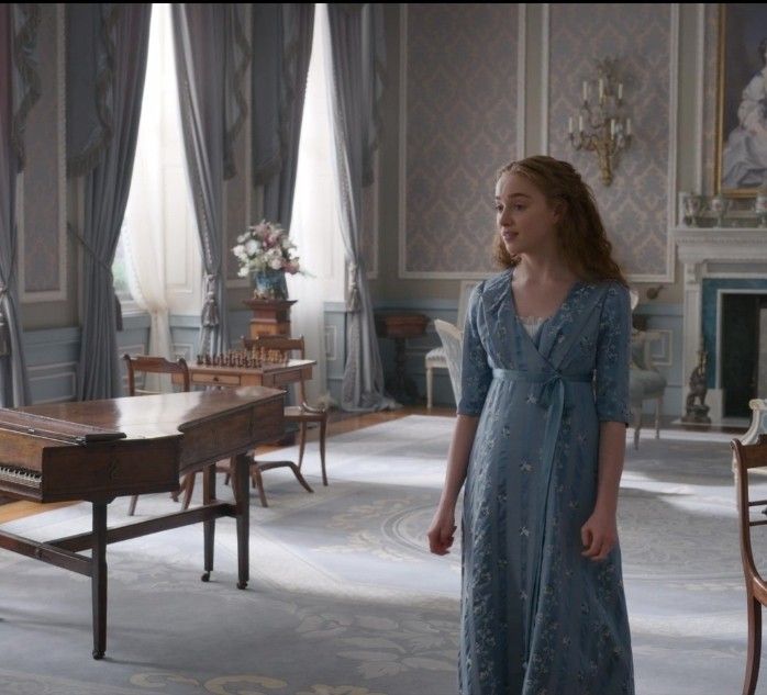 a woman in a blue dress standing in front of a table and chairs with a piano behind her