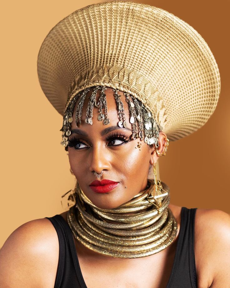 a woman wearing a large hat and gold jewelry on her neck, posing in front of a brown background