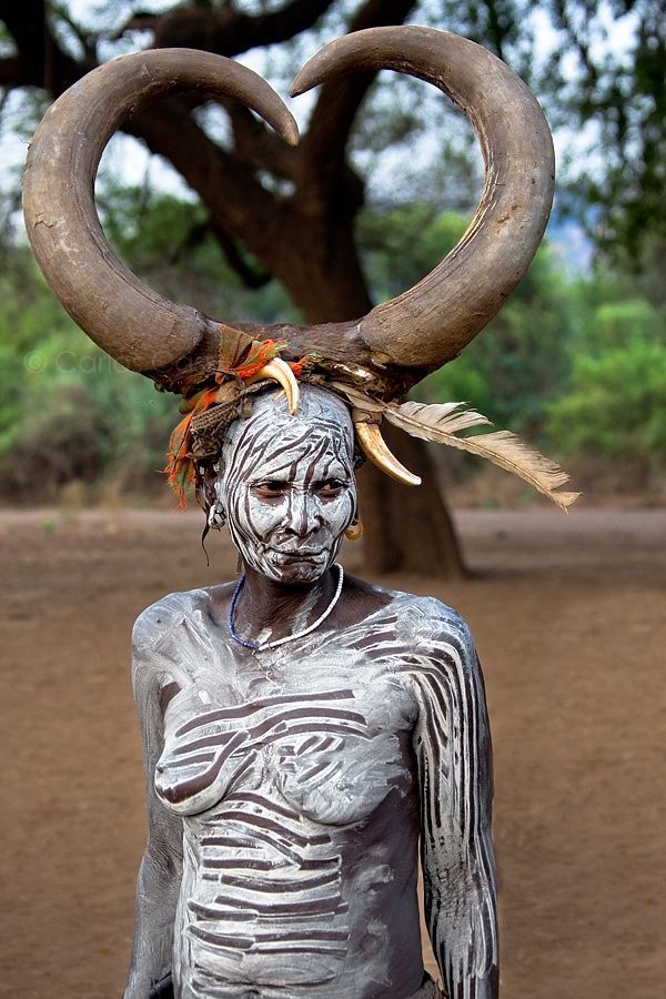 a woman with horns on her head standing in front of a tree and dirt ground